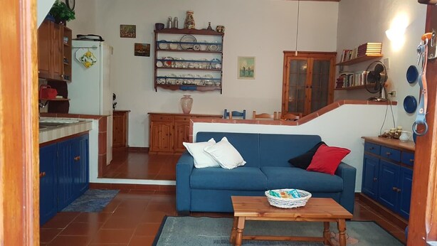 Una sala de estar con un sofá azul, una mesa de centro de madera, y una zona de cocina con armarios azules y piso de baldosas.