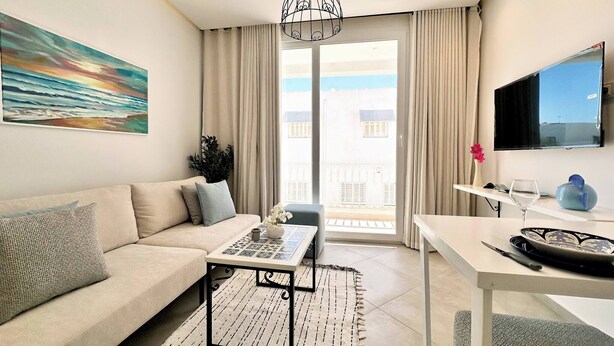A modern living room with a sofa, coffee table, and a dining table. There is a painting on the wall, a TV mounted on the wall, and a view of the outside through the window. - Appartement Nejma 2 Bedrooms (Sidi Bou Said)
