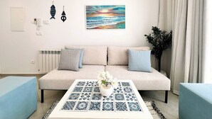 A living room with a sofa, a coffee table with a plant, a painting, and a radiator. - Appartement Nejma 2 Bedrooms (Sidi Bou Said)