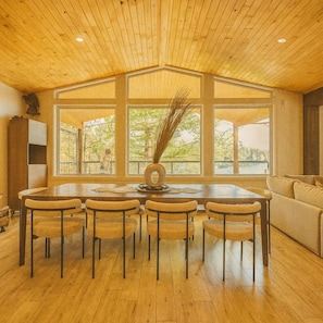 A dining area with a wooden table and chairs, large windows, and a view of the outdoors. - THE WOLF’S DEN - Modern Lakefront Cottage (Bancroft)