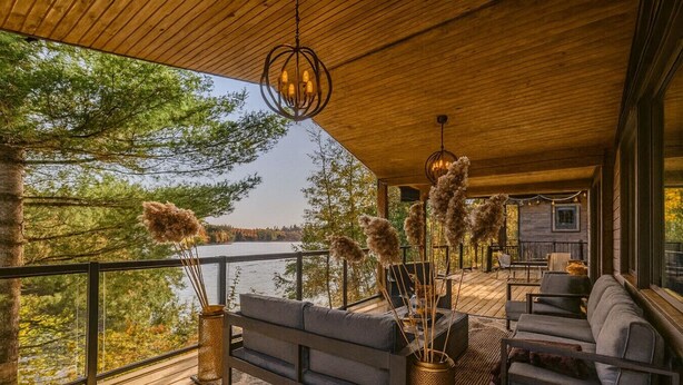 Eine Holzterrasse mit einer Couch und Stühlen, einem Geländer und Blick auf Bäume und Wasser.