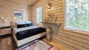 Ein Schlafzimmer einer Holzhütte mit Bett, Nachttisch, Lampe und einem Fenster mit Blick auf Bäume.