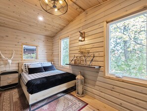 A wooden cabin bedroom with a bed, nightstand, lamp, and a window with a view of trees. - THE WOLF’S DEN - Modern Lakefront Cottage (Bancroft)