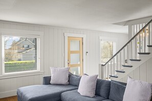 A modern living room with a grey sectional sofa, a wooden door, and a staircase with a metal railing. - 3BR 2BA Flamingos West (Seattle)