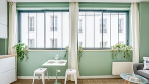 A room with a dining table, two stools, a sofa, a glass coffee table, and a view of buildings through large windows.