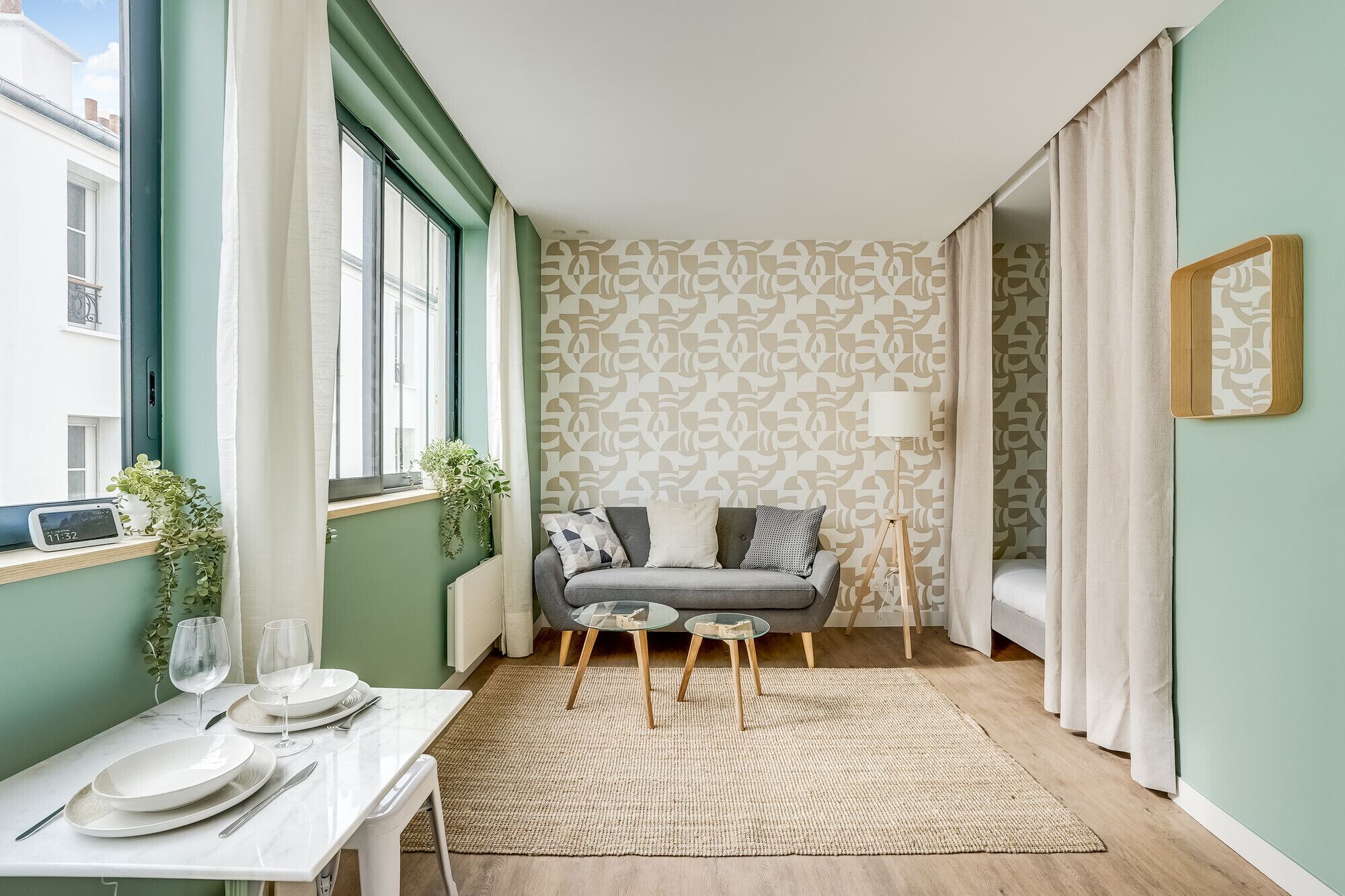 Et værelse med en sofa, et lille bord med en lampe, et vindue med gardiner og en væg med tapet.