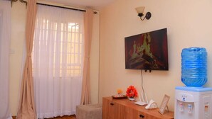 Een kamer met een aan de muur bevestigde televisie, een waterdispenser, een kastje met een bloevas en een raam met dunne gordijnen.