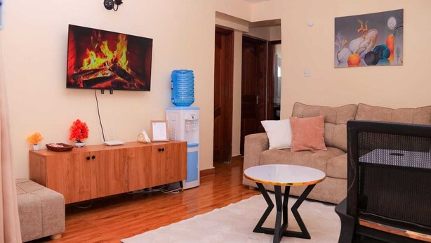 Een woonkamer met een bank, een salontafel, een televisietoestel waarop een haard te zien is, een waterdispenser en een schilderij aan de muur.