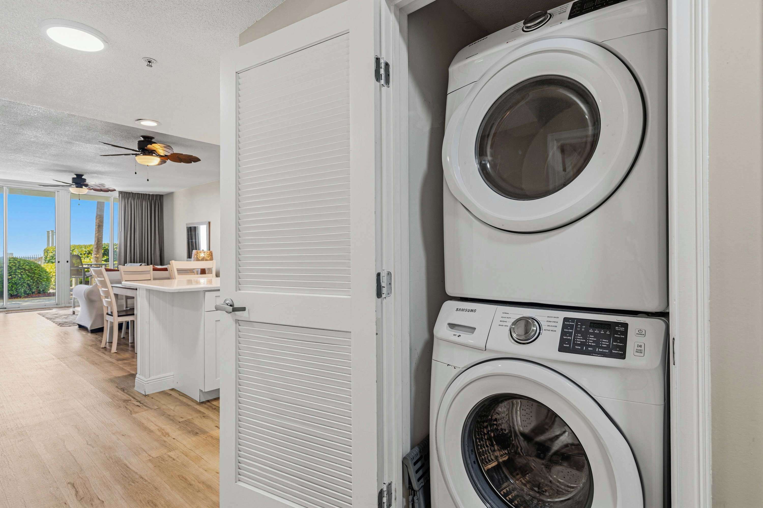 A laundry room with two stacked washing machines, a door, and a view of a dining area with chairs and a table.