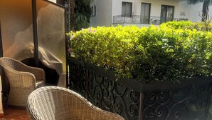A wicker chair, a small round table, a tiled floor, a decorative metal fence, and a building with balconies in the background. - Top location in Brissago 2 room apartment with private beach access. Dogs allowed (Brissago)