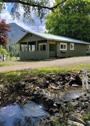 A rustic cabin with a porch, a small stream in front, and a mountain in the background. - Lake view, Private farm, King bed, Salmo/Fruitvale (Fruitvale)