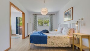 Makuuhuone, jossa on sänky, yöpöytä, riippuvalaisin, ikkuna verhoilla ja ovi, joka johtaa toiseen huoneeseen.