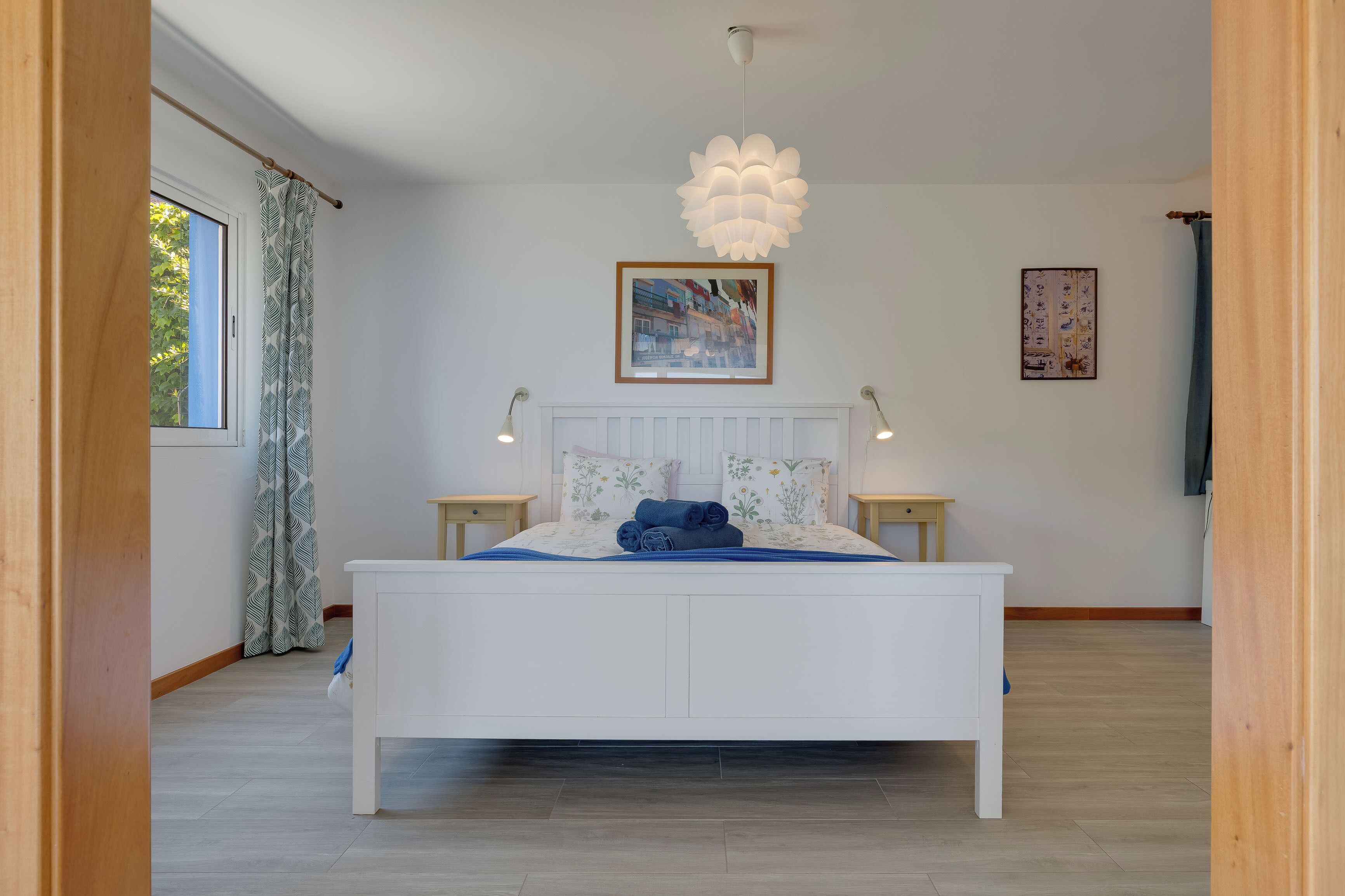 Een slaapkamer met een wit bed, twee nachtkastjes, een hanglamp, een raam met gordijnen en een ingelijst schilderij aan de muur.