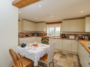 A kitchen with white cabinets, a dining table with a white tablecloth, and a window with a view of greenery. - Stanbridge Cottage (Malmesbury)