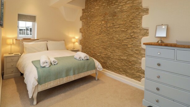 Een slaapkamer met een accentmuur van steen, een bed met witte lakens, twee nachtlampjes, een dressoir en een raam met uitzicht.