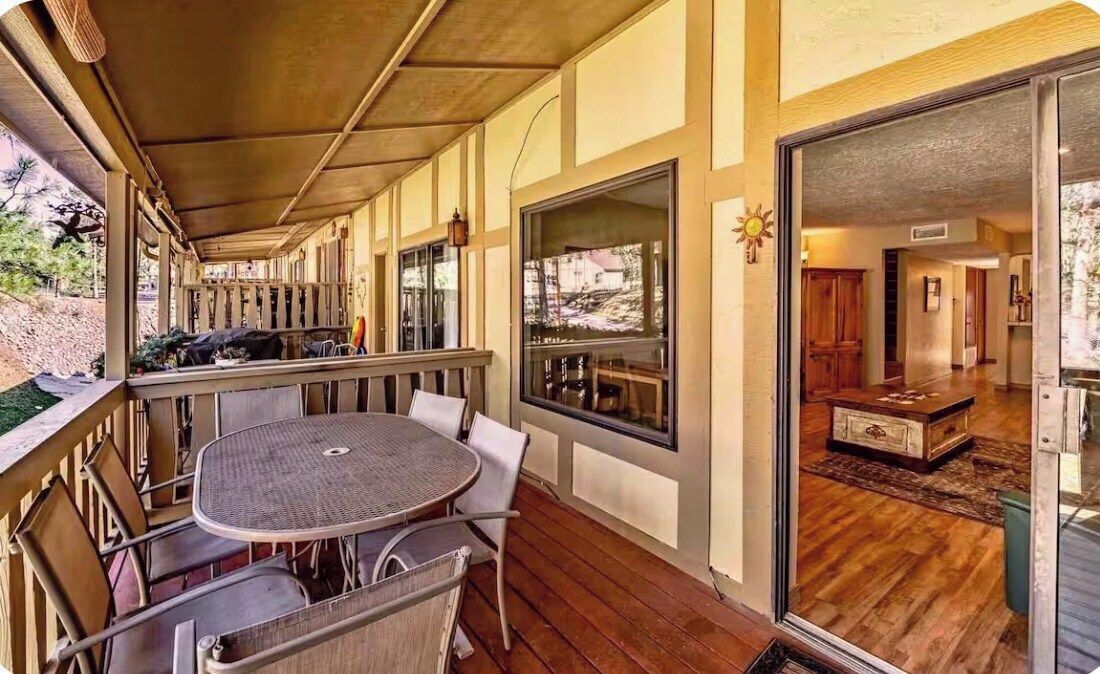 A covered outdoor patio with a round table and chairs, large windows, and a view of the interior.