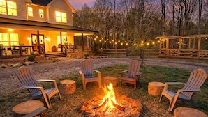 Een vuurkorf omringd door Adirondack-stoelen, een houten terras en een gebouw met verlichting op de achtergrond.