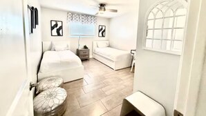 Un dormitorio compacto con cama, una pequeña zona de estar, ventilador de techo y una ventana con diseño de cuadrícula.