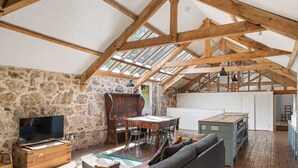 A loft-style room with a stone wall, wooden beams, a dining table, a sofa, and a television. - Hideaway on Dartmoor, Uphill Coach House (Lustleigh)