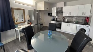 Eine kompakte Küche mit Kühlschrank, Mikrowelle, Herd und Waschmaschine. Ein Esstisch mit Stühlen und ein Glastisch sind ebenfalls vorhanden.