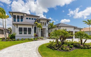A modern two-story house with a paved driveway, surrounded by well-maintained landscaping and palm trees. - New! Luxury Cape Coral Villa w/ Pool, Spa & Dock (Cape Coral)
