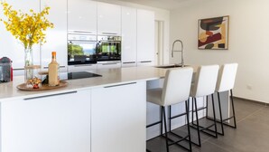 A modern kitchen with white cabinetry, a central island with bar stools, a built-in oven, and a sink. - Amara 4 - Luxury villa with sea view (Les Trois-Îlets)