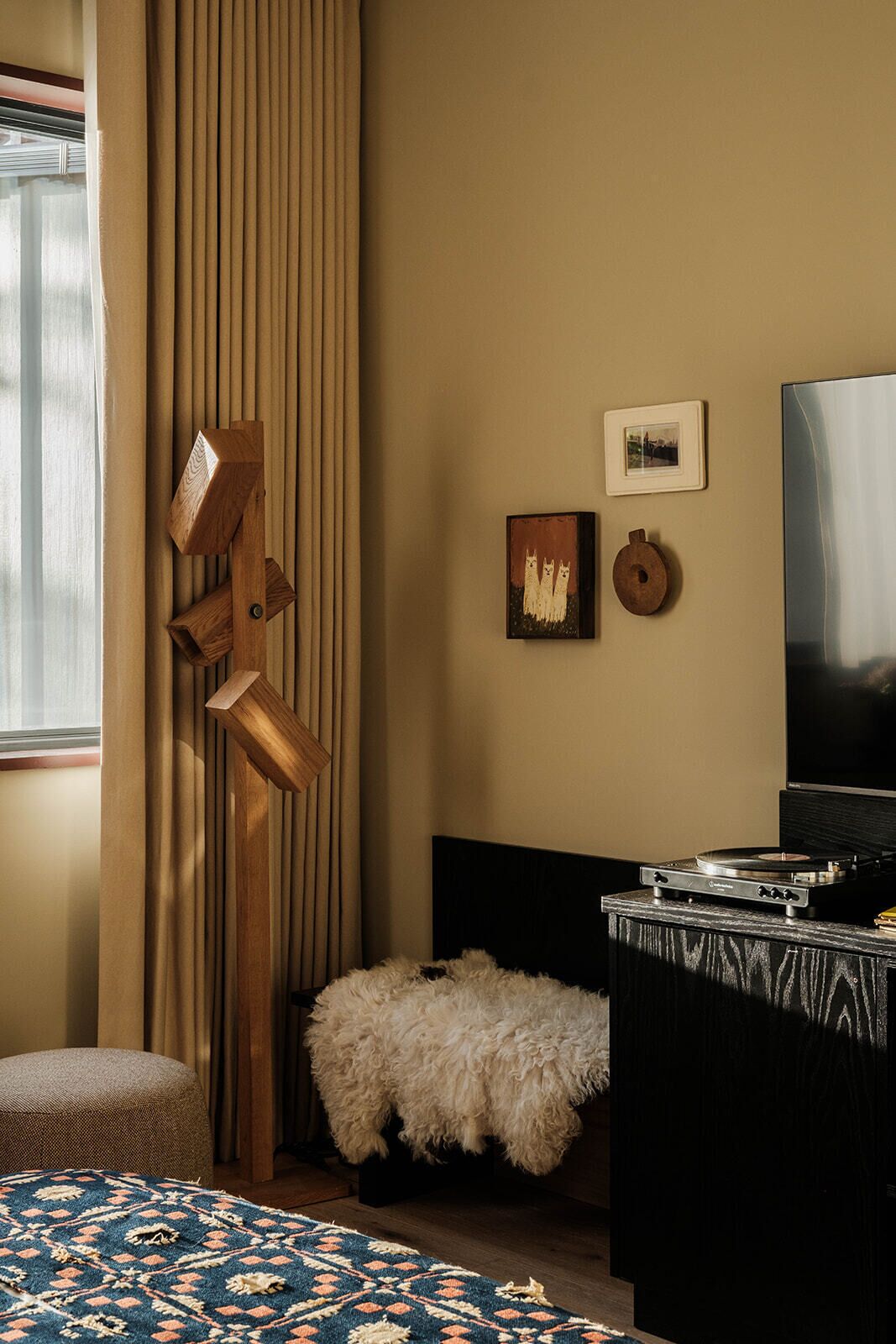 Una habitación con una cama, un televisor, un estante de madera montado en la pared, un otomano blanco mullido y una alfombra estampada.