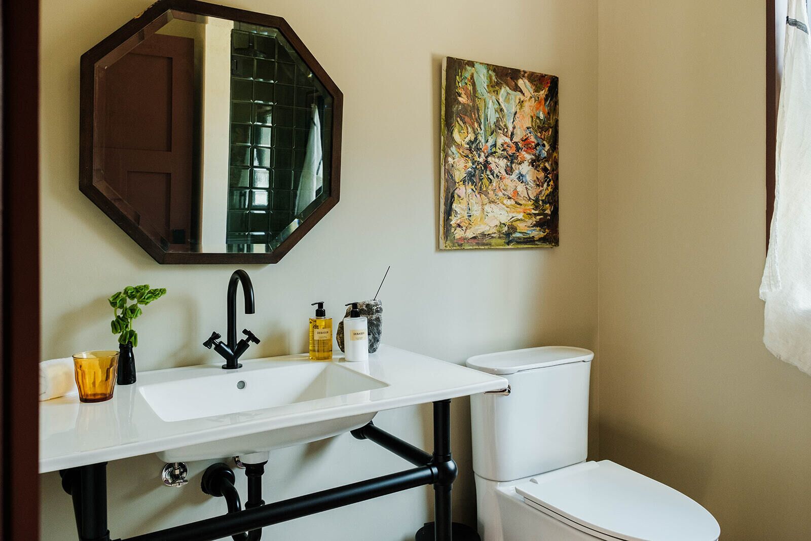 A bathroom with a white sink, black faucet, and a toilet. There is a mirror above the sink and a painting on the wall.