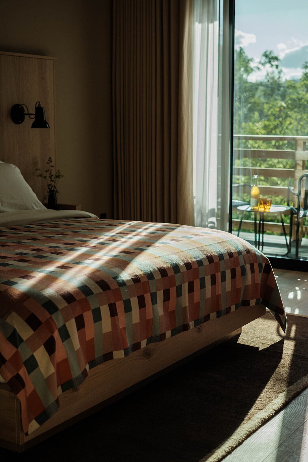 A bedroom with a large bed, a nightstand, a lamp, a window with curtains, and a view of greenery outside.