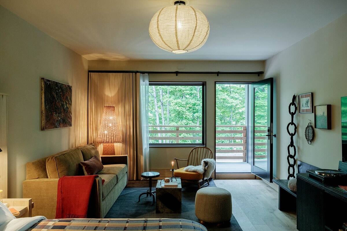 A cozy living room with a sofa, a chair, a small table, and a lamp. There are large windows with a view of greenery outside.