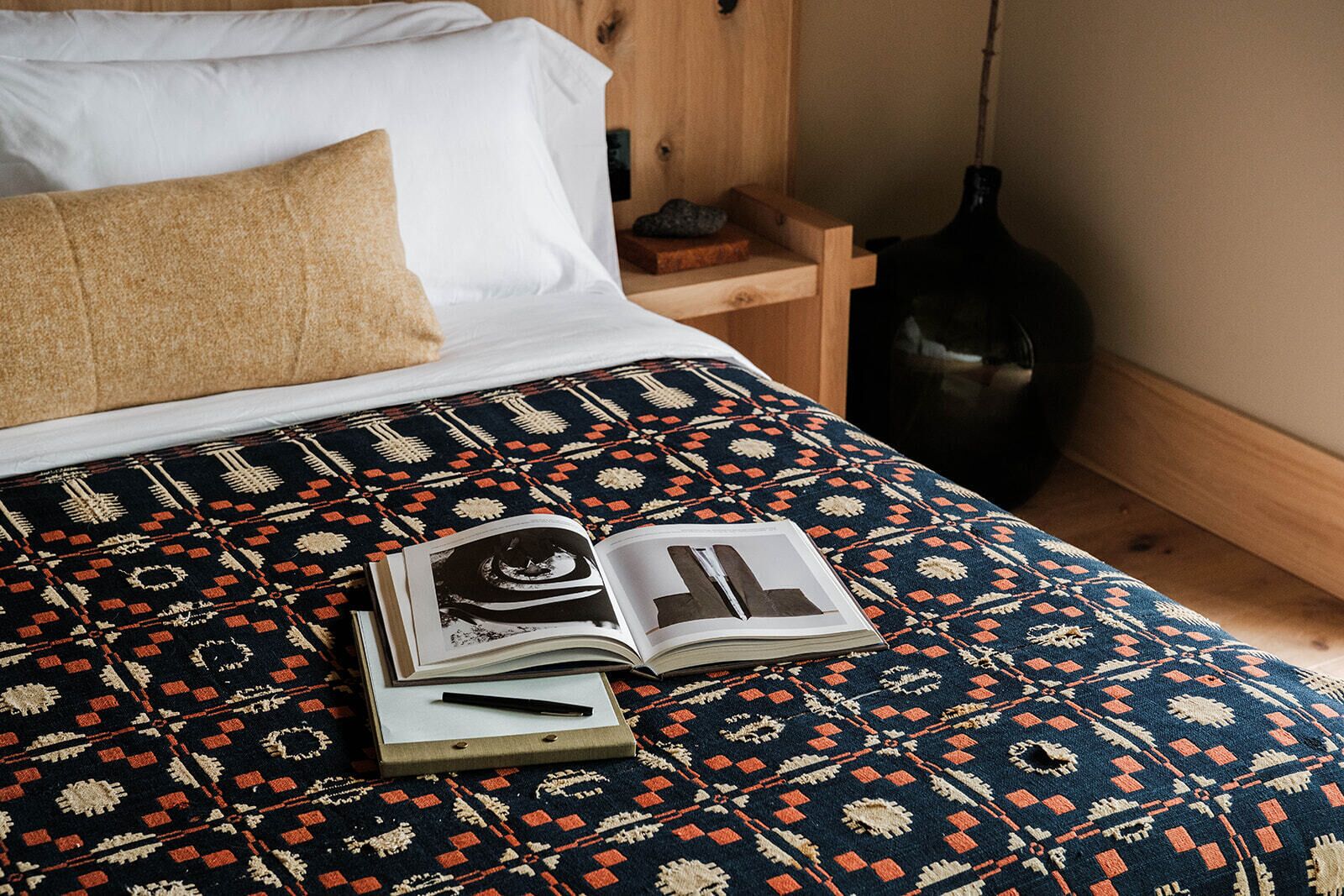 A bed with a patterned bedspread, a book, and a pen on it.