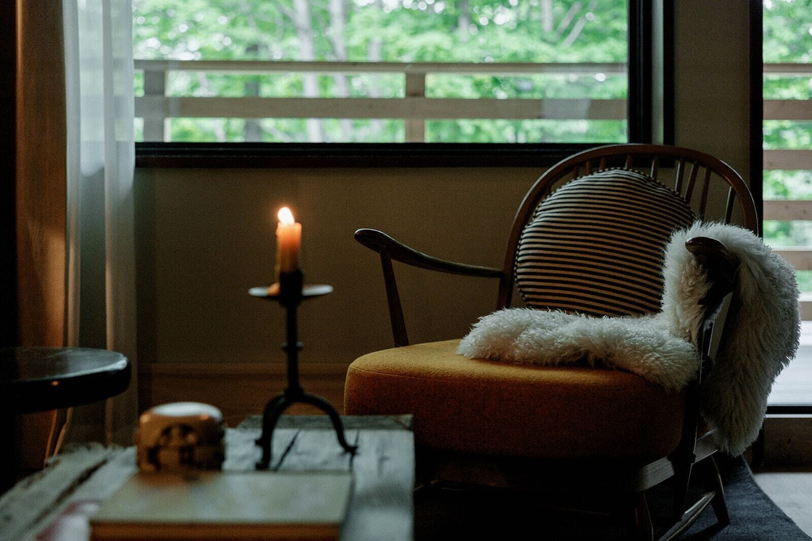 A chair with a fluffy pillow, a lit candle on a stand, and a window with a view of greenery.