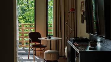 A room with a wooden chair, a small round table, a red floor lamp, a television, and a view of greenery outside.