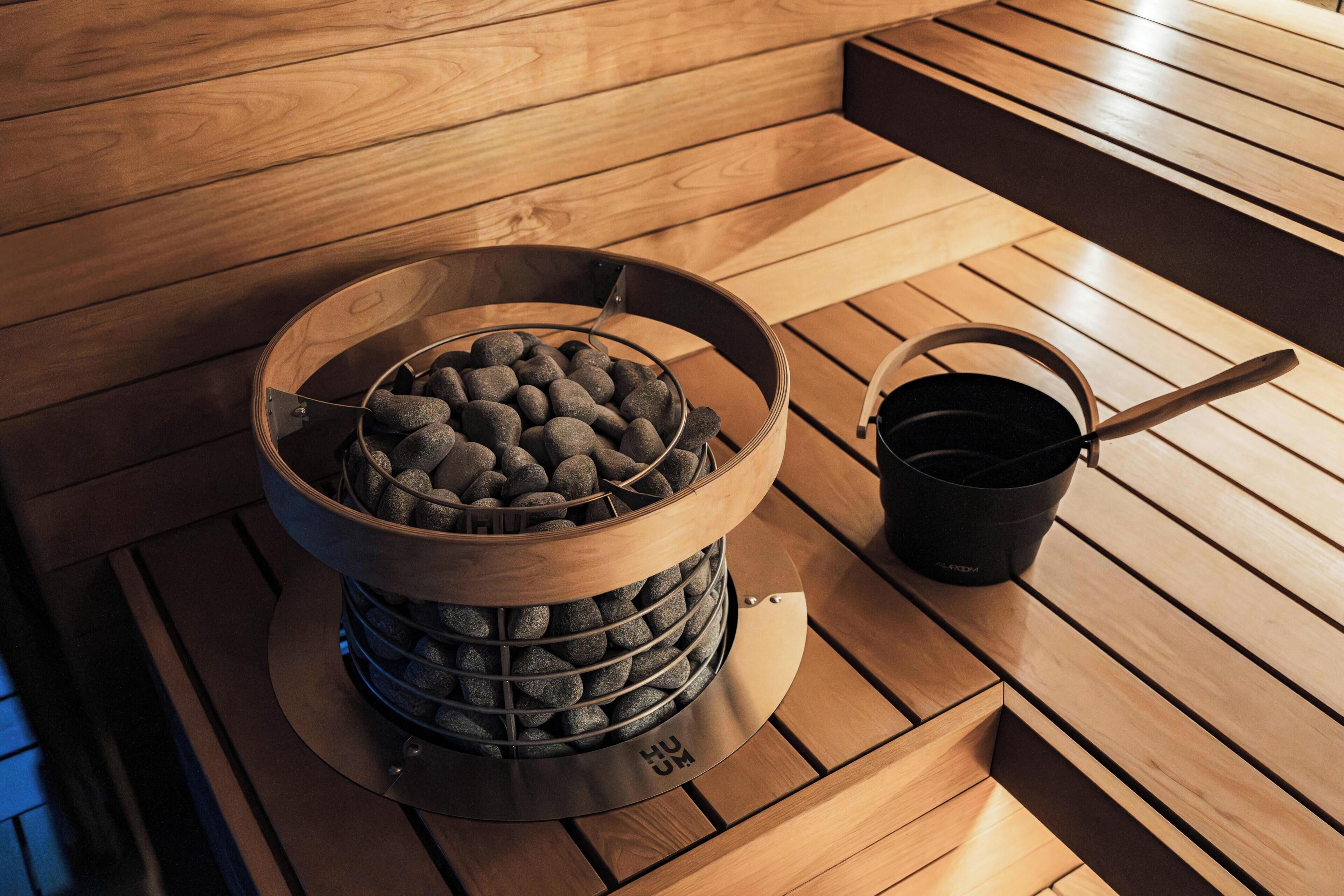 A sauna with a wooden bench, a metal container with stones, and a black bucket with a handle.