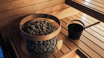 A sauna with a wooden bench, a metal container with stones, and a black bucket with a handle.