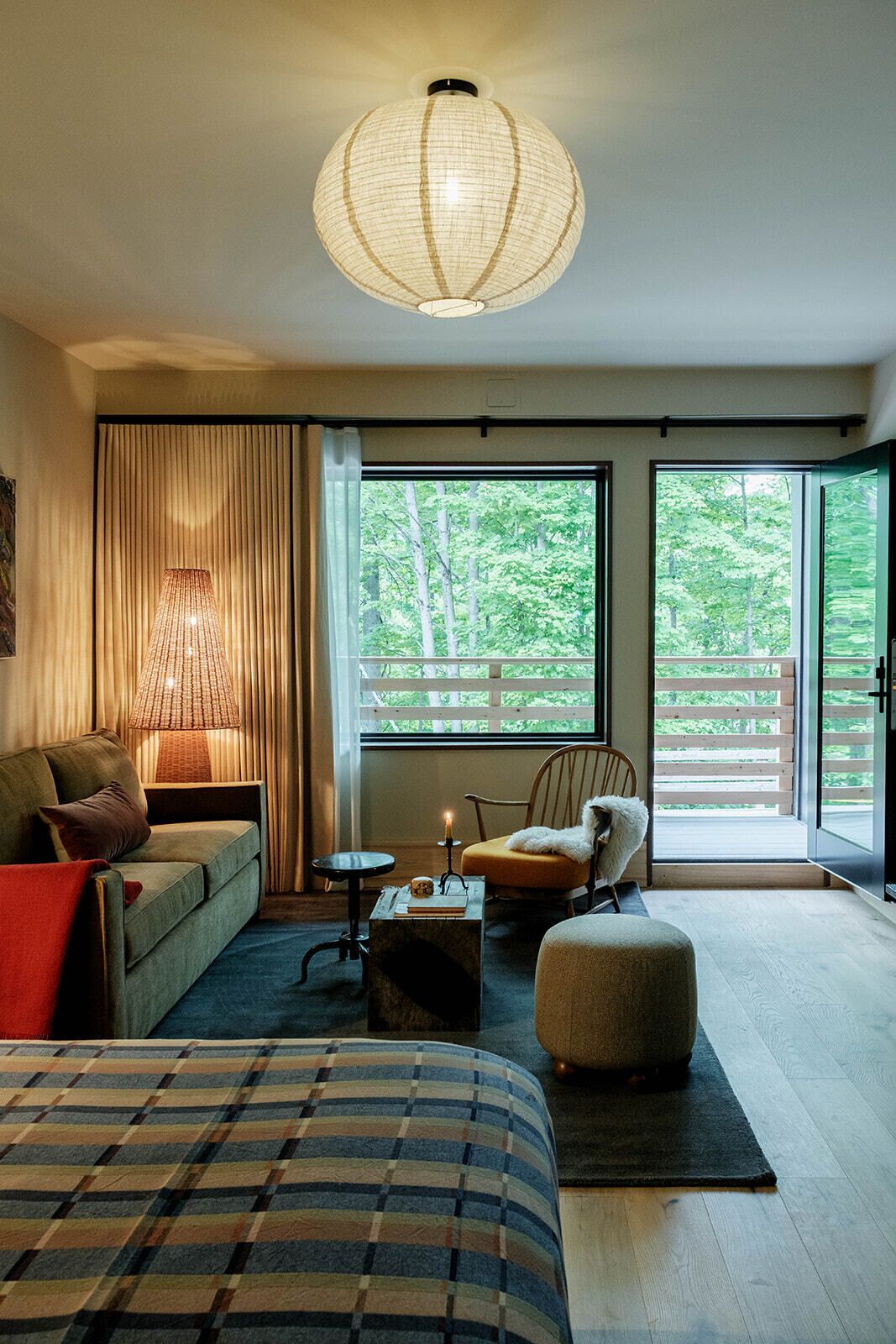 A modern bedroom with a large bed, a sofa, a chair, a small table, and a large window offering a view of trees.