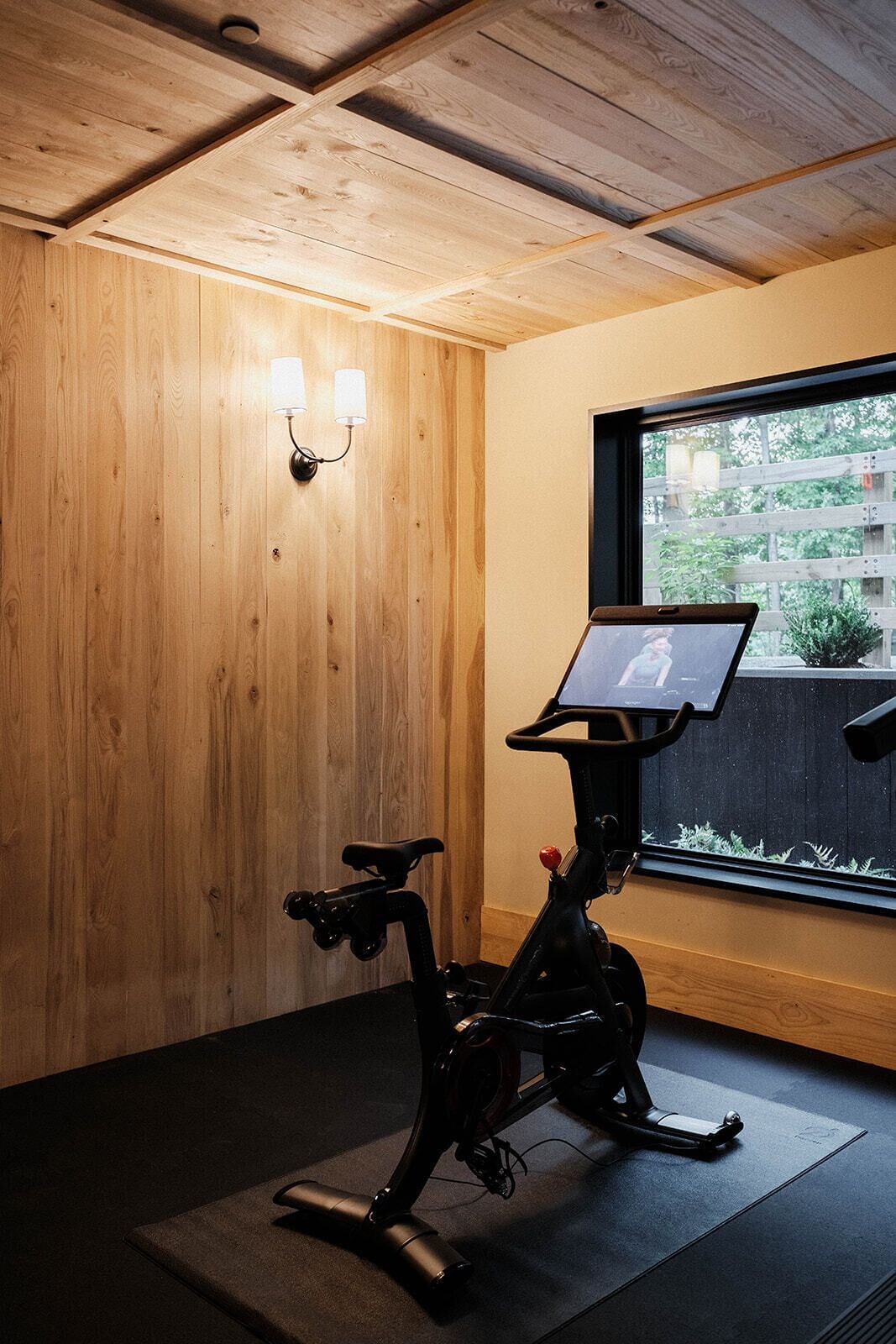 A stationary bike with a screen, a wall-mounted lamp, and a window with a view of greenery.