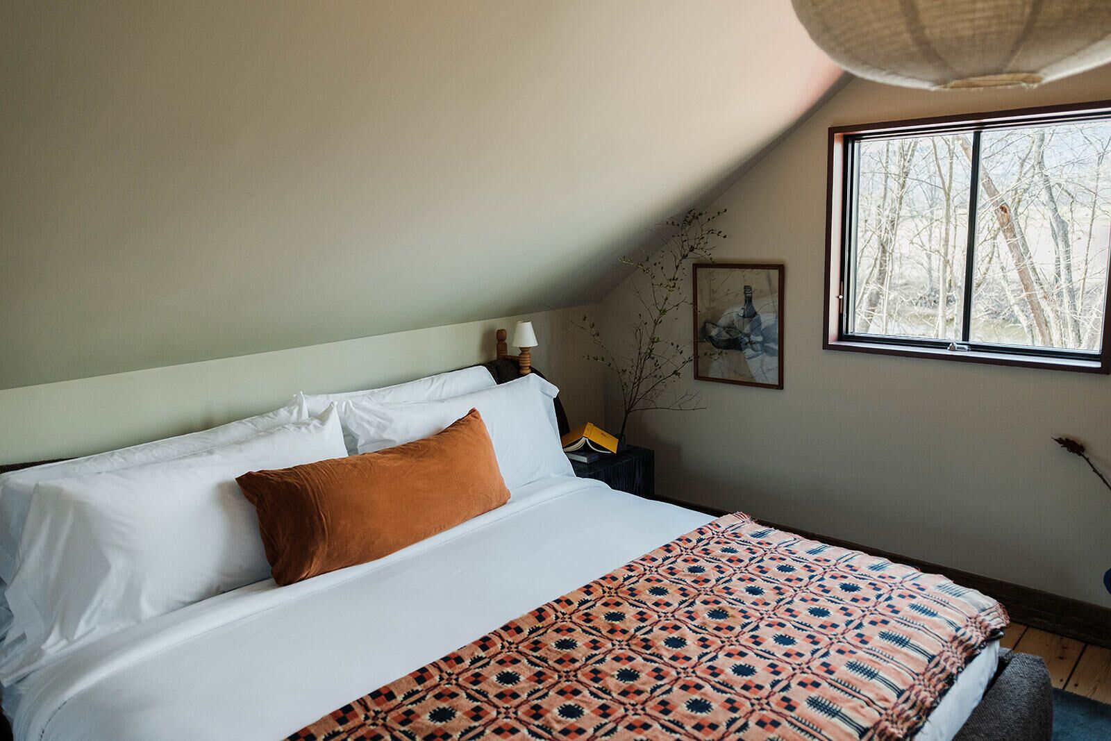 A neatly made bed with white and brown bedding, a patterned throw, and a window with a view of bare trees.
