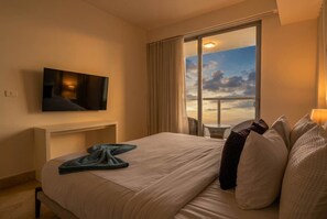 Ein Hotelzimmer mit Bett, einem an der Wand befestigten Fernseher, einem Balkon mit Vorhängen und Blick auf den Himmel.