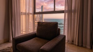 Ein Zimmer mit Meerblick und einem hohen Gebäude, ein brauner Sessel und durchsichtige Vorhänge.