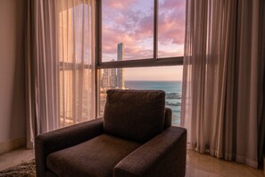 A room with a view of the ocean and a tall building, a brown armchair, and sheer curtains. - 20B Apartamento de 1 recamara, con vista al mar (Panamá)