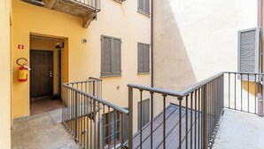 Un escalier étroit avec des rampes métalliques menant à une porte, un extincteur fixé au mur, et un bâtiment à la façade beige et aux volets.