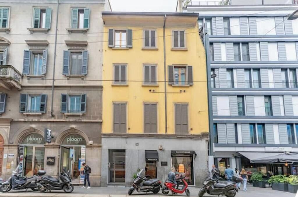 Straßenansicht mit Gebäuden, darunter ein gelbes Gebäude mit einer Schaufenstereingangspartei mit der Aufschrift 'ploom' und ein graues Gebäude mit einer Schaufenstereingangspartei mit der Aufschrift 'ploom'.