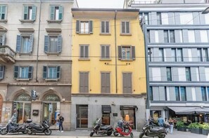 A street view with buildings, including a yellow building with a storefront labeled 'ploom' and a gray building with a storefront labeled 'ploom.' - AP6 Apartment in the Cathedral area (Milano)