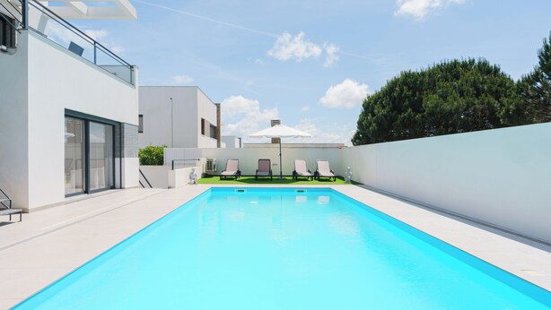 Una piscina rectangular con superficie azul cristalina, rodeada por un muro blanco y un edificio moderno con grandes ventanales y un balcón.