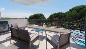 Un balcón con mobiliario de mimbre, una mesa de vidrio y vistas a árboles y edificios.