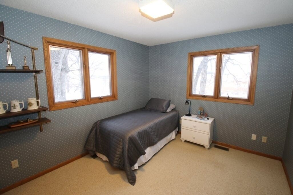 A bedroom with two windows, a bed, a nightstand, a shelf with items, and a light fixture.