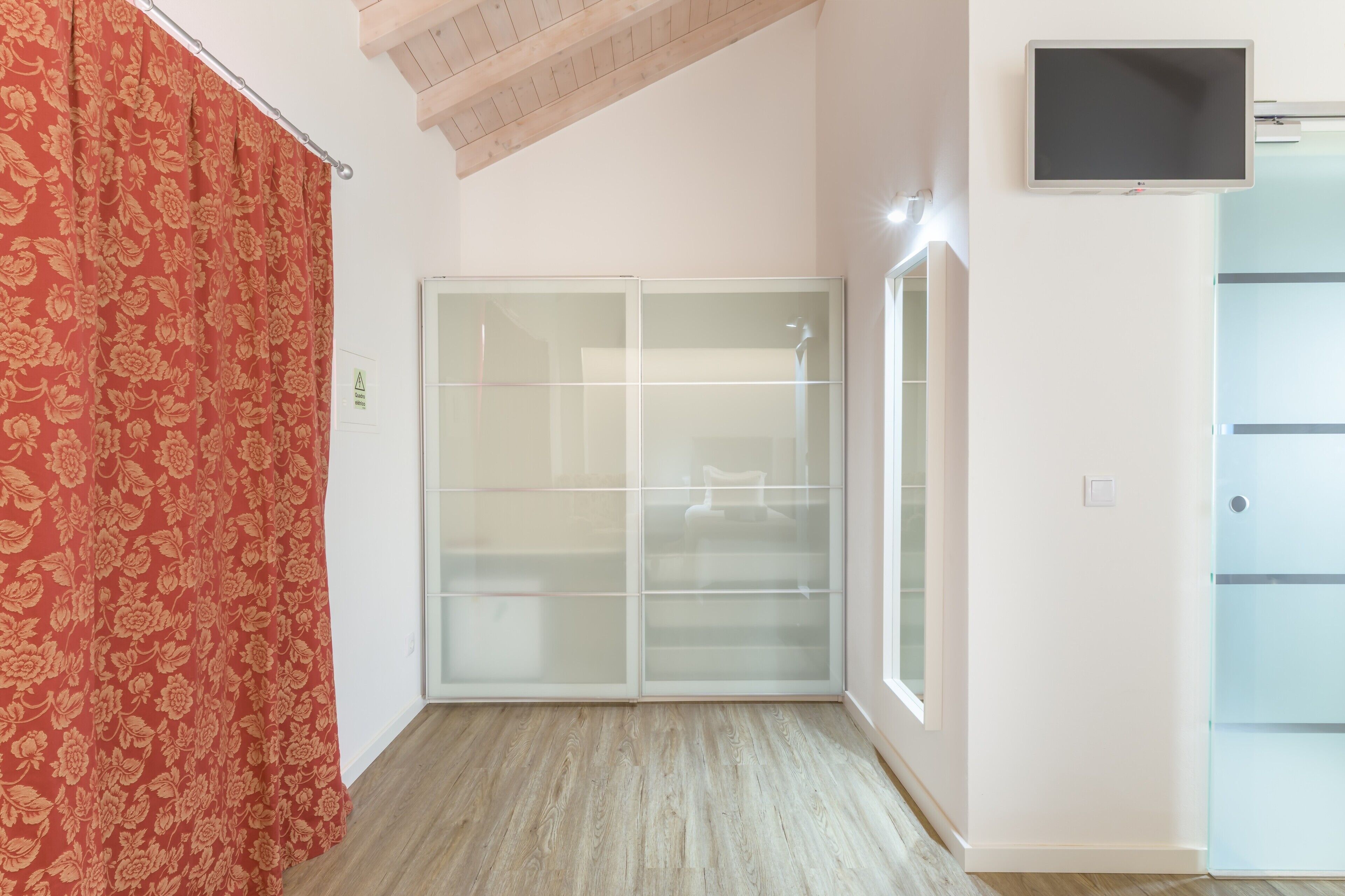 A room with a sliding glass door closet, a flat-screen TV mounted on the wall, and a wooden ceiling.