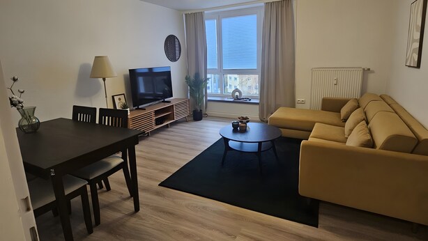 Moderni olohuone, jossa on ruokapöytä, sohva, televisio ja suuri ikkuna.