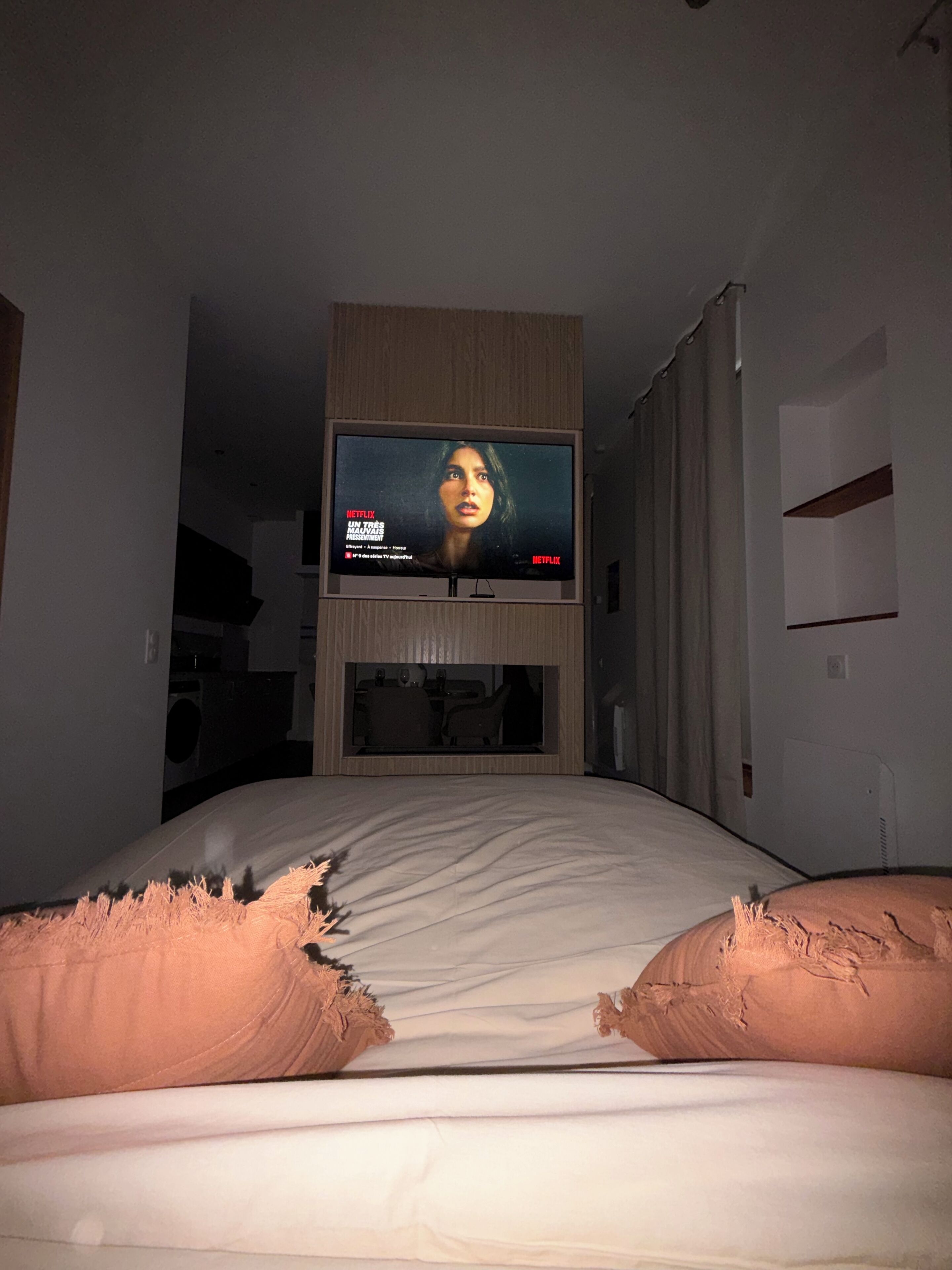 Une chambre dotée d’un grand téléviseur à écran plat fixé au mur, d’un lit avec une literie blanche et d’une tête de lit en bois.